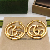 Gold earrings with interlocking 'G' design on a beige cloth with a green box in the background.